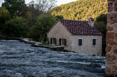 Hırvatistan 'ın Krka ulusal parkında çağlayan şelalelerin kenarında duran geleneksel su değirmeni binasının manzarası, doğa ve mimarinin pitoresk bir manzarasını oluşturuyor.
