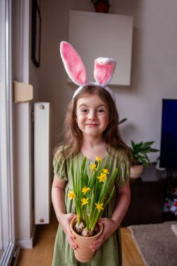 Tavşan kulağı takan gülümseyen küçük kız elinde çiçek açan nergislerle bir terasotta tenceresi tutuyor, Paskalyayı ve baharın gelişini evinde kutluyor.