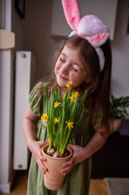Sevimli küçük bir kız tavşan kulağı takıyor, terasotta saksısını tutuyor ve kokluyor. Çiçek açan nergislerin tadını çıkarıyor, baharın tadını çıkarıyor ve Paskalyayı kutluyor.