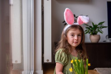 Paskalya bayramını kutlayan sevimli bir çocuk, tavşan kulaklığı takan ve neşeli bir buket nergis tutan neşeli bir şekilde şenlikli süslemelerle dolu oturma odasında gülümseyen.