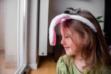 Tavşan kulaklığı takmış gülümseyen küçük bir kız Paskalya kutlamalarının neşeli beklentisiyle pencereden dışarı bakıyor çocukluk mutluluğunun ve şenlik ruhunun özünü yakalıyor.