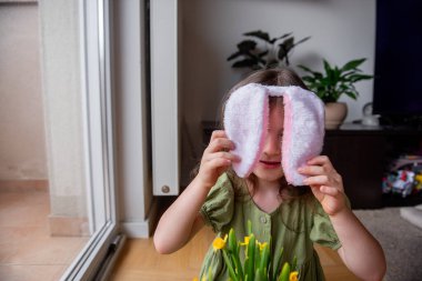 Paskalya tavşanı kulaklarını önünde tutan küçük bir kız, neşeli bir oyun oynuyor ve neşeli Paskalya sezonunda eğleniyor, çiçek açmış nergislerle çevrili.