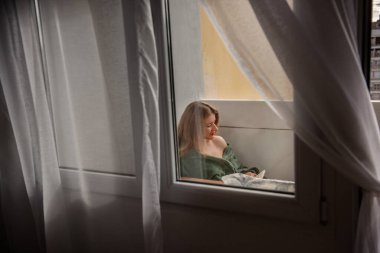 Yeşil ekose elbiseli kızıl saçlı kadın balkonuna oturmuş, sessiz ve düşünceli bir anın tadını çıkarırken bir deftere bir şeyler yazıyor. Yumuşak perdeler, huzurlu açık hava sahnesini çerçeveler..