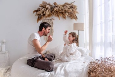 Baba ve kızı, parlak beyaz bir yatak odasında rahat bir yatakta oturup oyun oynuyor ve sabah ışığında neşeli anlar paylaşıyor, kalıcı anılar yaratıyorlar.