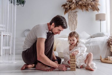 Baba ve kızı yatak odasında yerde oturuyor, dikkatle ahşap bloklardan bir kule inşa ediyorlar, neşeli anları paylaşıyorlar ve eğlenceli etkileşim ve konsantrasyonla bağlarını güçlendiriyorlar.