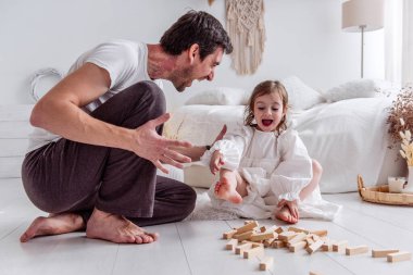 Babası ve küçük kızı yerde oturup neşe içinde ahşap bloklarla oynuyorlar. Kahkahalarını paylaşıyorlar ve samimi yatak odalarında güzel anılar yaratıyorlar.