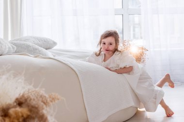 Beyaz bir elbise giyen sevimli küçük bir kız beyaz bir yatak odasında neşeyle oynuyor, neşeli bir sabah havasını kucaklıyor.