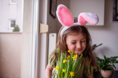 Tavşan kulaklığı takan sevimli küçük kız evde bir buket taze nergisin kokusunu alıyor, Paskalya atmosferinin tadını çıkarıyor ve baharın gelişini kutluyor.