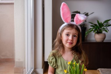 Paskalya tavşanı kulakları takan sevimli anaokulu kızı, evinde neşeli bir portre için poz verirken neşe dolu bir buket nergis tutuyor ve mutluluk saçıyor.
