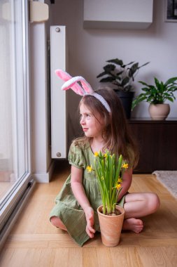 Paskalya tavşanı kulaklığı takan şirin bir çocuk elinde sarı nergislerle pencerenin yanında oturmuş Paskalya tavşanını bekliyor.