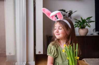 Küçük kız tavşan kulağı takıyor, elinde neşe dolu sarı nergis buketi, uzaklara bakarken neşeyle gülümsüyor, Paskalya bayramını evinde kutluyor.