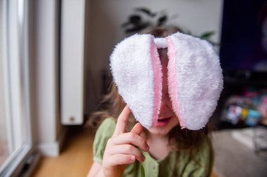 Küçük kız tavşan kulaklarıyla oynayarak eğleniyor gözlerini kapatıyor, çocukluk oyunları, Paskalya tatili konsepti, yeşil bir elbise giyiyor ve evinin içinde.