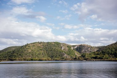 Visovac gölü, güneşli bir günde Hırvatistan 'ın Krka ulusal parkında güzel bir manzara olan yemyeşil bitki örtüsü ve tepelerle çevrili sakin sularıyla bulutlu gökyüzünü yansıtıyor.