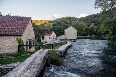 Krka Nehri 'nin temiz suları Hırvatistan' ın Krka ulusal parkındaki yemyeşil ağaçlar ve tepelerle çevrili döşemeli çatıları olan eski taş evlerin yanından akıyor