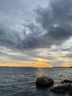 Koyu bulutların ardında batan altın güneş, sakin bir denizin üzerine sıcak ışınlar saçıyor. Ön plandaki kayalar sahneyi güzelleştiriyor, alacakaranlıkta çarpıcı ama sakin bir atmosfer yaratıyor.