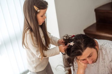 Genç bir kız, babasının saçını renkli klipslerle şekillendiriyor, sıcak bir ev ortamında ebeveynler ve çocuklar arasında neşeli ve bağ kuran bir an sergiliyor.