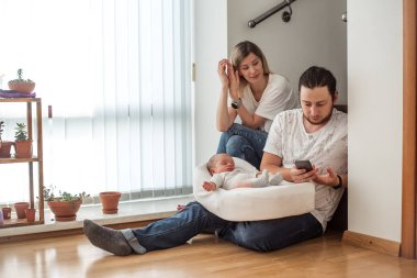 Genç ebeveynler yeni doğmuş bebeklerine bakmak için evde oturuyorlar, anne onlara göz kulak olurken baba telefonunu kullanıyor.