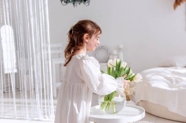 Beyaz elbiseli şirin küçük kız beyaz masadaki cam bir vazoda bir buket beyaz lale düzenliyor. Parlak odayı barışçıl ve masum bir atmosferle dolduruyor.