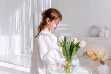 Gecelikli, parlak, güneşli bir odada beyaz laleleri düzenleyen, çiçek bakımının huzurlu anının tadını çıkaran ve bahar tazeliğinin güzelliğini kucaklayan sevimli bir çocuk. Boho iç mimarisi