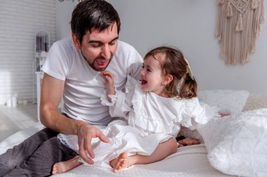 Baba ve kızı beyaz bir yatak örtüsünde oturuyor, aynı beyaz pijamaları giyiyorlar, kahkahaları ve neşeli anları paylaşıyorlar, sıcak ve neşe dolu bir yatak odasında.
