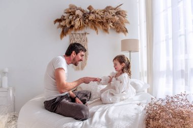 Baba ve kızı bir yatakta oturuyor, taş kağıt makas oynuyorlar, sıcacık bir bohem yatak odasında, kurumuş çiçekler ve makrome ile güzel vakit geçiriyorlar.