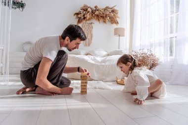 Baba ve kızı yatak odasında yerde oturuyor, dikkatle ahşap bloklardan bir kule inşa ediyorlar, neşeli anları paylaşıyorlar ve eğlenceli etkileşim ve konsantrasyonla bağlarını güçlendiriyorlar.