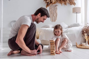 Baba ve kızı yatak odasında yerde oturuyor, dikkatle ahşap bloklardan bir kule inşa ediyorlar, neşeli anları paylaşıyorlar ve eğlenceli etkileşim ve konsantrasyonla bağlarını güçlendiriyorlar.