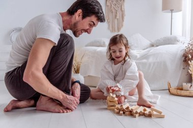Babası ve küçük kızı yerde oturup neşe içinde ahşap bloklarla oynuyorlar. Kahkahalarını paylaşıyorlar ve samimi yatak odalarında güzel anılar yaratıyorlar.
