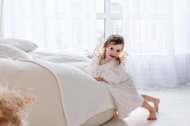 Beyaz bir elbise giyen sevimli küçük bir kız beyaz bir yatak odasında neşeyle oynuyor, neşeli bir sabah havasını kucaklıyor.