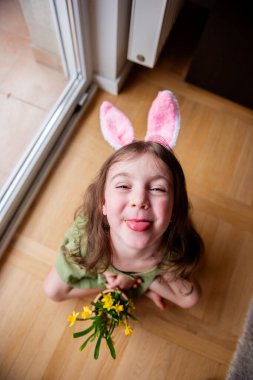 Pembe tavşan kulaklı küçük kız, tahta zeminde otururken ve elinde sarı nergis saksısı tutarken dilini dışarı çıkarıyor.