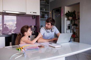Baba evde çalışıyor, mutfak masasında genç kızına online okul ödevlerinde yardım ediyor, destekleyici ve birlikte öğrenme ortamı sağlıyor.