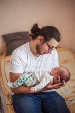 Genç baba bir yatakta oturuyor ve uyuyan yeni doğmuş bebeğini kucağında tutuyor. Sevgi, ilgi ve şefkat dolu bir aile anı yaşıyor.