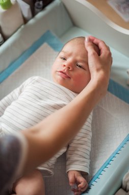 Anne yeni doğmuş bebeğini nazikçe soyunma masasında yatarken okşar. Çizgili tulum giyen bebek, annesinin nazik dokunuşunun altında sakin ve hoşnut görünüyor.
