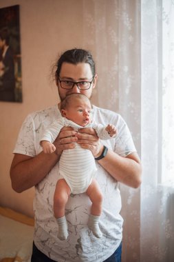 Uzun saçlı ve gözlüklü baba bebeğini sıcak bir evde tutuyor. Bebek çizgili bir tulum giyiyor ve doğal ışık perdeden akıyor.