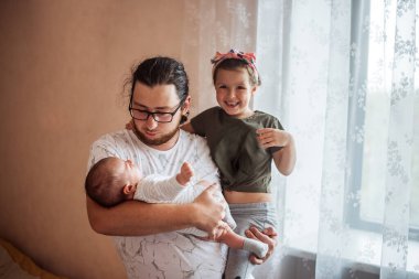 Babası bebeğini sevgiyle tutarken neşeli kızı yanında duruyor. Sıcak, sade atmosfer, parlak bir odada aile bağını ve mutluluğu vurguluyor.