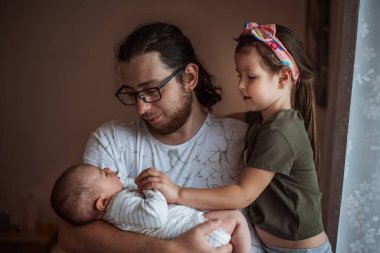 Genç baba yeni doğmuş bebeğini kucaklarken kızı da ona şefkatle bakıyor. Evinde sevgi ve bağla dolu güzel bir aile portresi yakalıyor.