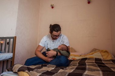 Genç, uzun saçlı, gözlük takan ve günlük giysiler giyen bir baba. Yeni doğmuş bebeğini battaniyeye sarmış, beşiğine sarılmış, sevgi ve özen gösteren bir şekilde yatakta oturuyor.