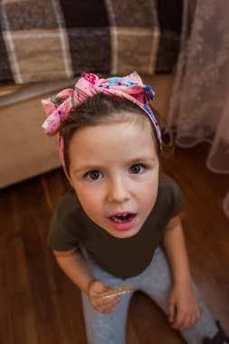 Renkli saç bandı takan genç bir kız, şaşırmış bir ifade sergiliyor, ahşap bir zeminde oturuyor. Ayarlar yumuşak döşenmiş rahat bir iç mekan içeriyor