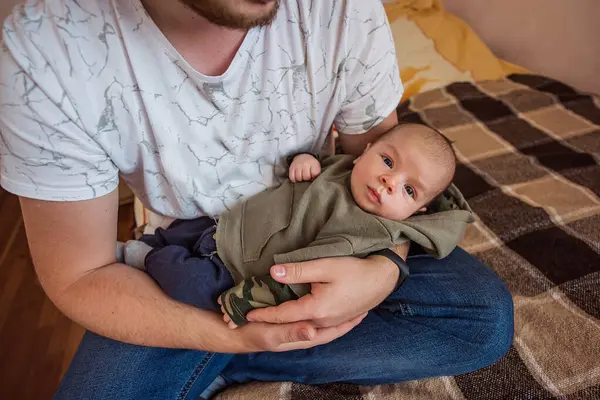Baba sevgiyle kucağında yeni doğmuş bebeğini tutuyor ve sıcak ve samimi bir an yaratıyor. Bebek rahat bir kıyafete sarılmış, rahatça babanın korumasına sokulmuş.