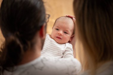 Ebeveynler sevgi dolu bir şekilde yeni doğan bebeklerini kucaklarken ailenin sıcaklığını ve bağını yakalıyorlar. Bebek, çizgili bir kıyafet giymiş, yukarı bakıyor, masumiyet ve huzur taşıyor.