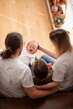 Ebeveynler ağlayan bebeklerini yerde otururken rahatlatıyorlar, yeni yürümeye başlayan kızlarını yakın tutuyorlar. Sıcak, besleyici bir ev ortamında sevgi ve destek anları