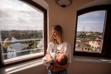 Genç anne, yeni doğmuş bebeğini emzirirken evinin penceresinin yanında durmuş, doğal ışığın ve şehrin manzarasının tadını çıkarıyor.