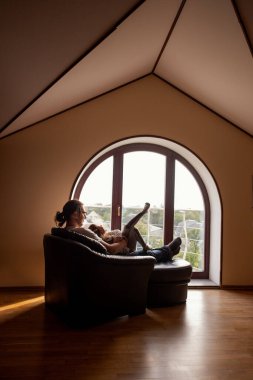 Büyük kemerli bir pencerenin yanında rahat bir sandalyede uzanan bir adam, dışarıdaki huzurlu manzaranın tadını çıkarıyor. Sıcak güneş ışığı tavan arasını dolduruyor. Huzurlu ve davetkar bir atmosfer yaratıyor.