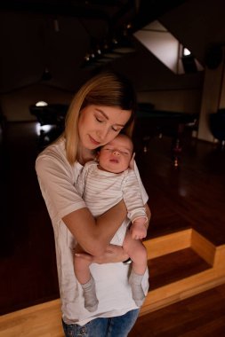 Huzurlu bir an, uyuyan bebeğini sıcak ve rahat bir evde nazikçe tutan bir anneyi yakalıyor. Ahşap zemin ve yumuşak ışıklandırma huzurlu bir atmosfer yaratır.