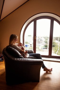 Anne, bebeğini rahat deri bir sandalyede otururken sevgiyle kucaklıyor. Geniş kemerli pencere, huzurlu bir açık hava manzarası sunuyor, huzurlu ve samimi bir atmosfer yaratıyor.