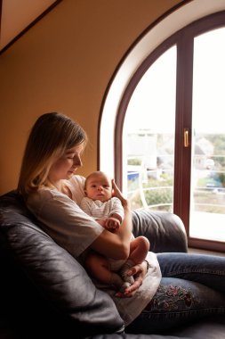 Genç anne bir kanepede oturuyor ve yeni doğmuş bebeğini kollarında tutuyor, evlerinin geniş kemerli penceresinin yanında, sıcak ve samimi bir annelik ve aile bağı sahnesi yaratıyor.