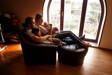 Genç bir aile rahat bir koltukta birlikte vakit geçirmenin keyfini çıkarıyor, geniş kemerli bir pencereden dışarı bakıyor, doğal ışıkta güneşleniyor ve aşk ve birliktelik anlarını paylaşıyor.