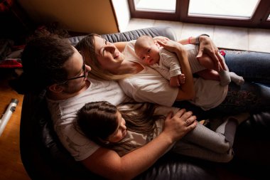 Ebeveynler yeni doğmuş bebekleri ve kızlarıyla rahat bir kanepede uzanmış evde huzurlu bir anın tadını çıkarıyorlar. Huzurlu bir atmosferde sevgi ve beraberliği somutlaştırıyorlar.