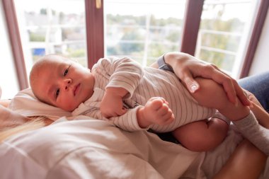 Bir annenin kucağında rahat bir şekilde uzanan bir bebeğin huzurlu sahnesi. Pencereden süzülen yumuşak doğal ışık. O an sıcaklığı, şefkati ve aile sevgisini yakalar.