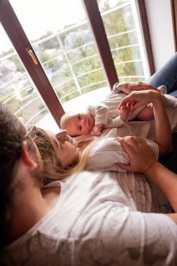 Pencerenin kenarında yatan, yeni doğan bebeklerine sarılan ve kucaklayan genç ebeveynler sevgi ve bağla dolu huzurlu ve samimi bir aile anı yaratıyorlar.